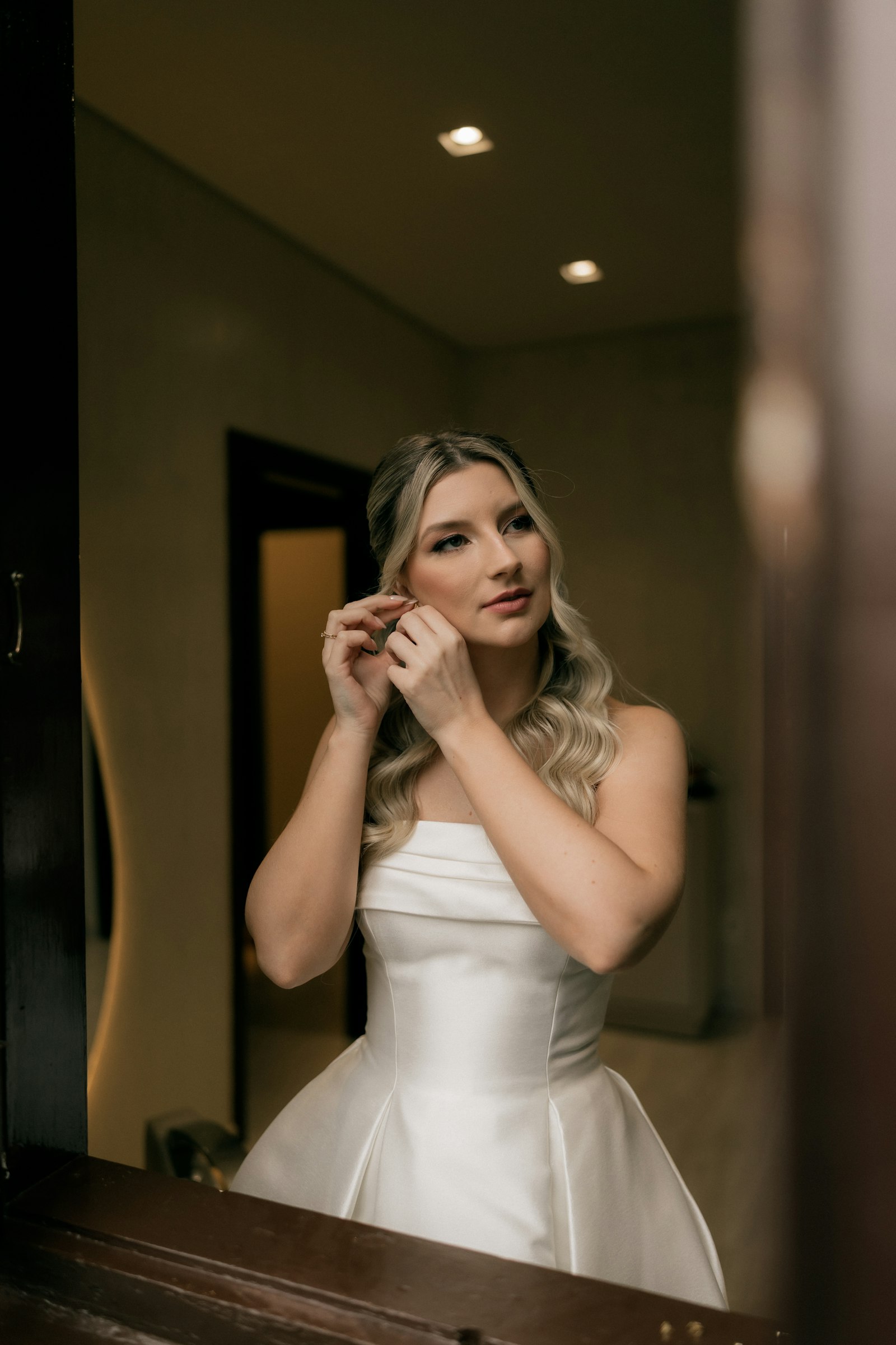 Noiva de vestido branco colocando o brinco em frente ao espelho