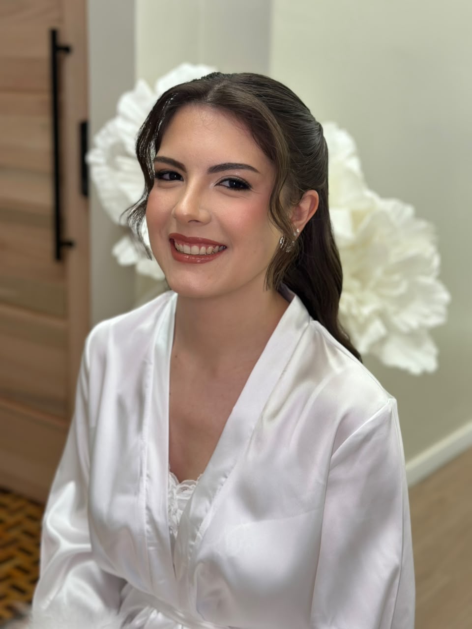 Noiva sorridente de robe branco, com penteado semipreso e flores brancas ao fundo